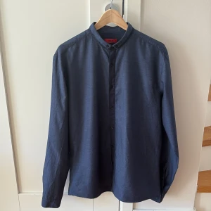Mörkblå skjorta från Hugo Boss - Säljer en stilren mörkblå skjorta från Hugo Boss i slim fit-modell. Skjortan har ståkrage, knäppning framtill och långa ärmar. Perfekt för dig som gillar en klassisk men modern look. Står XL som storlek men passar mer som L.