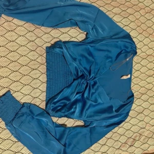 Blå croppad blus från Amisu - Säljer en blå croppad blus från Amisu i glansigt material. Toppen har långa ärmar med breda muddar och smockad midja. Perfekt för dig som vill ha en trendig och snygg topp med lite extra detaljer.