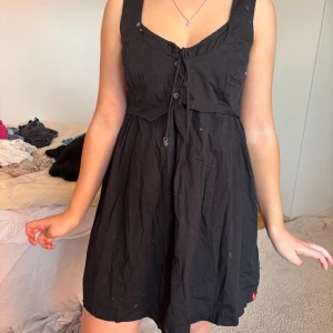 Svart kortklänning med knytning - Säljer en svart kortklänning med knytning framtill och breda axelband. Klänningen har en lätt utställd passform och är perfekt för varma dagar. Enkel och stilren design som passar till många tillfällen.