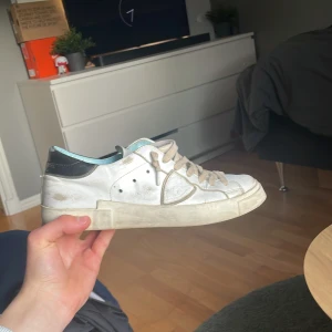 Philippe Model skor - Säljer ett par vita Philippe Model sneakers med svarta detaljer på hälen och ljusblå insida. Skorna har snörning och en platt sula. Klassisk och stilren design med rund tå och diskreta sömmar. Skorna är storlek 40