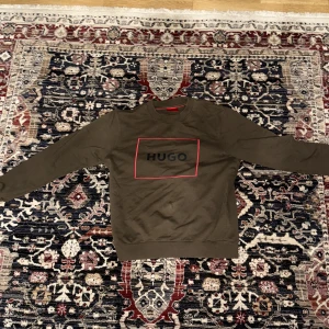 Olivgrön sweatshirt från Hugo - Säljer en olivgrön sweatshirt från Hugo med rund halsringning och röd ram runt loggan på bröstet. Tröjan har långa ärmar och en klassisk passform. Perfekt för en stilren och avslappnad look.