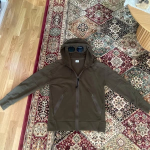 C.P. Company hoodie med goggles  - Säljer en olivgrön hoodie från C.P. Company med ikoniska inbyggda glasögon i huvan. Jackan har dragkedja framtill, två snedställda fickor med dragkedja och justerbar huva med snörning. Perfekt för dig som gillar streetwear och unika detaljer.