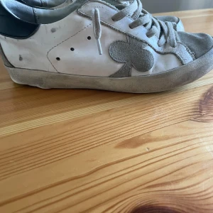 Vita sneakers med grå mockadetaljer - Snygga vita sneakers med grå mockadetaljer och svart häl. Skorna har snörning och en unik klubb-symbol i mocka på sidan. Ovandelen är i skinn och mocka, sulan är svart och slitstark. Perfekta för dig som gillar en stilren men ändå unik look. Obs dom är limade därav priset 