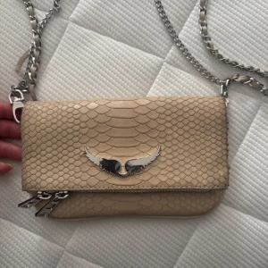 Beige axelväska med kedjerem och ormskinnsmönster - Säljer en beige axelväska från zadig med snyggt ormskinnsmönster och silverfärgad kedjerem. Väskan har dubbla dragkedjor och en ikonisk silverfärgad fågeldetalj på framsidan. Perfekt för dig som vill ha en stilren accessoar med det lilla extra. Den har lite defekter på sidan och kedjan, visas på bilderna.