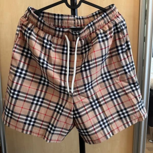 Beige rutiga shorts från Burberry - Snygga beige shorts från Burberry med klassiskt rutigt mönster i svart, vitt och rött. Shortsen har resår i midjan och vit snörning för justerbar passform. Perfekta för sommaren och har en exklusiv känsla. Stl L och passar folk mellan 170-185