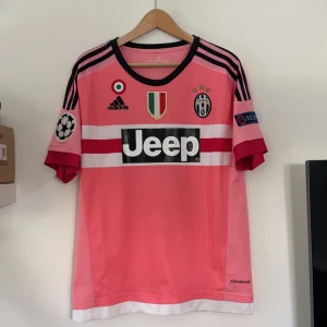 Juventus rosa bortatröja Dybala 21 - Juventus bortatröja i rosa med svarta och vita detaljer, Adidas-märke och Jeep sponsortryck. Tröjan har Dybala 21 på ryggen, Champions League och Respect-patchar på ärmarna samt klubbmärke och tre stjärnor på bröstet.