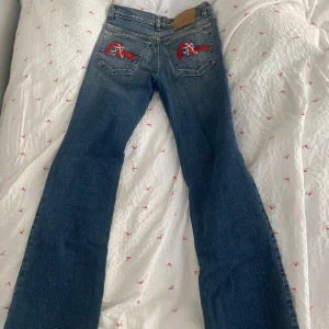 Blå lågmidjade jeans från Evisu med broderade bakfickor - lågmidjade blå jeans från Evisu med broderade röda och vita detaljer på bakfickorna. Midjemått: 35cm Innerbenslängd: 82/83cm
