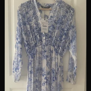 Blommig långklänning från Zara - Säljer en långklänning från Zara med blått och vitt blommönster. Klänningen har v-ringning och långa ärmar, samt en lätt rynkad midja för en feminin siluett. Perfekt för sommarens tillfällen.