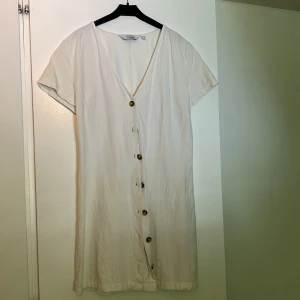 Vit klänning med knappar från & Other Stories - Säljer en vit klänning från & Other Stories med v-ringning och knappar framtill. Klänningen har korta ärmar och ett knytband i midjan för en snygg passform. Perfekt för varma dagar och enkel att styla.