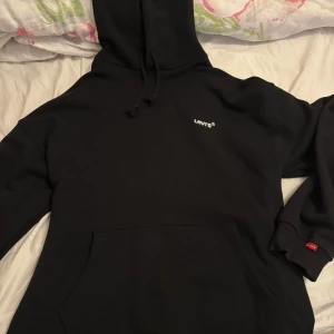 Svart hoodie från Levi's - Säljer en svart hoodie från Levi's med liten vit logga på bröstet och klassisk känguruficka framtill. Tröjan har huva med dragsko och ribbade muddar vid ärmslut. Perfekt för en avslappnad stil.