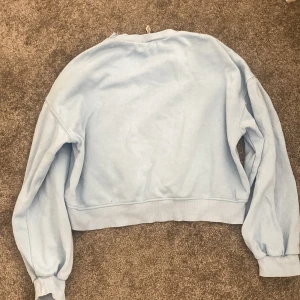 Ljusblå croppad sweatshirt - Säljer en ljusblå croppad sweatshirt med långa ärmar och bred mudd i midjan. Perfekt för en avslappnad och trendig stil. Enkel design utan tryck eller mönster.