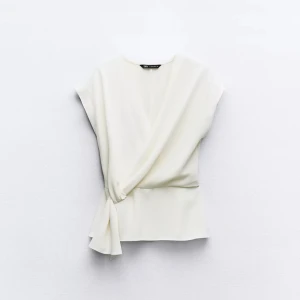 Vit omlottopp från Zara - Säljer en stilren vit omlottopp från Zara med knyt i sidan. Toppen har korta ärmar och en elegant v-ringning. Perfekt för dig som gillar minimalistisk och fräsch stil.