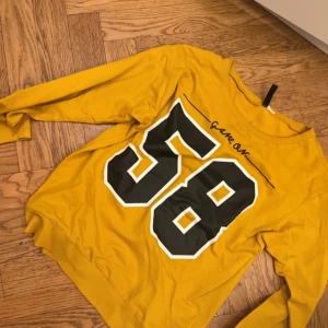 Gul sweatshirt med tryck - Gul sweatshirt med stort svart och vitt tryck av siffran 58 på framsidan och texten 'Game On' vid halsen. Tröjan har långa ärmar och en avslappnad passform. Perfekt för en sportig look.