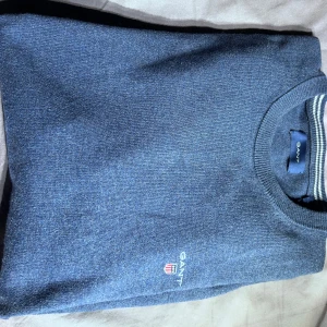 Blå tröja från GANT - Säljer en klassisk blå tröja från GANT med rund halsringning och diskret broderad logga på bröstet. Tröjan har ribbade muddar och en stilren look som passar perfekt till jeans eller chinos.