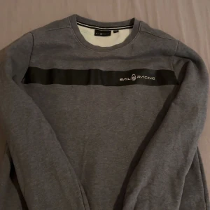 Grå sweatshirt från Sail Racing - Grå sweatshirt från Sail Racing med svart band och logga över bröstet. Rund halsringning och långa ärmar. Perfekt för en avslappnad stil och enkel att matcha med jeans eller joggers.