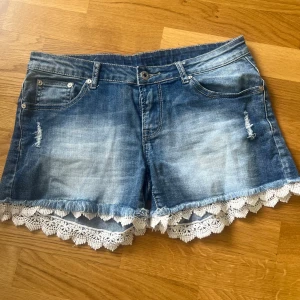 Blå jeansshorts med spetskant - Säljer ett par blå jeansshorts med snygg slitning och vit spetskant längst ner. Shortsen har fem fickor och klassisk knappgylf. Perfekta för sommaren och ger en söt touch till din outfit.