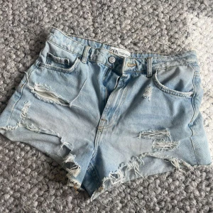 Ljusblå jeansshorts från Zara - Ljusblå jeansshorts från Zara med slitna detaljer och fransar. Klassisk femficksmodell med hög midja och råa kanter. Perfekta för varma dagar och en avslappnad stil.