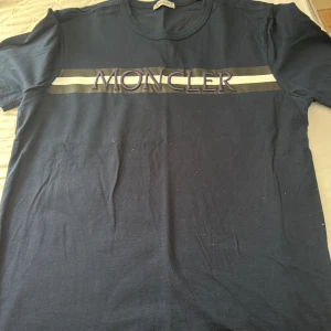 Mörkblå t-shirt från Moncler med broderad logga - Säljer en mörkblå t-shirt från Moncler med broderad logga och ränder över bröstet. Klassisk rund halsringning och korta ärmar. Perfekt för dig som gillar stilrena märkesplagg.storlek m men passar s