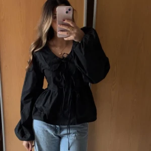 Svart blus med volang och knyt - Säljer en svart blus med v-ringning, volangdetalj vid halsen som man knyter framtill. Så fin passform då den går in lite i midjan och balongärmarna är även de en fin detalj!