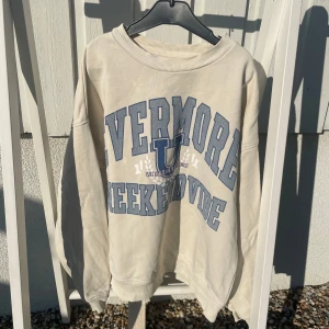 Beige collegetröja från Lindex med tryck - Säljer en beige collegetröja från Lindex med blått tryck på bröstet där det står 'VERMORE WEEKEND VIBE'. Tröjan har rund halsringning och långa ärmar. Perfekt för en avslappnad stil.