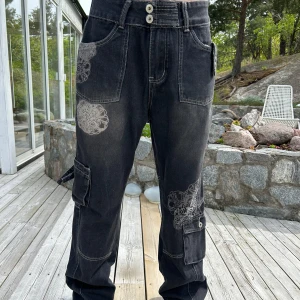 Svarta cargopants med broderade detaljer - Säljer ett par svarta cargopants med vita broderade mönster på benen. Byxorna har flera fickor, synliga sömmar och knappar i midjan. Perfekta för dig som gillar en lite tuffare stil med unika detaljer.🤗