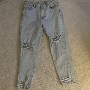 Ljusblå jeans med hål - Säljer ett par ljusblå jeans med slitningar på båda knäna och vid bensluten. Jeansen har klassisk femficksdesign och hög midja. Perfekta för en avslappnad och trendig look.