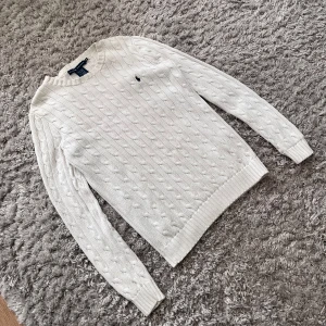 Vit kabelstickad tröja från Ralph Lauren - Säljer en klassisk vit kabelstickad tröja från Ralph Lauren i 100% bomull. Tröjan har lång ärm, rund halsringning och den ikoniska broderade loggan på bröstet. Perfekt för en stilren och tidlös look.