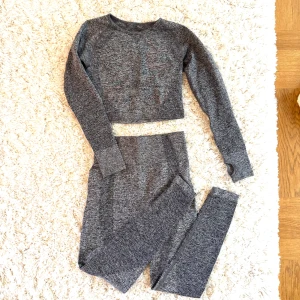 Tränings set - Säljer ett set med gråmelerad croppad långärmad träningströja och matchande tights. Tröjan har tumhål i ärmsluten och båda plaggen är i stretchigt material som passar perfekt till träning.