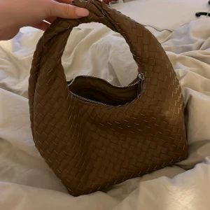 Brun flätad axelväska - Säljer en snygg brun axelväska med flätad design och dragkedja upptill. Väskan har en mjuk, rundad form och ett kort handtag som gör den enkel att bära på axeln. Perfekt accessoar för att lyfta din outfit.