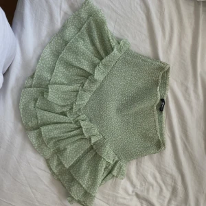 Ljusgrön volangkjol från Gina Tricot - Supersöt ljusgrön kjol från Gina Tricot med volanger och diskret mönster. Kjolen har en kort passform och är perfekt för vår och sommar. Resår i midjan för extra komfort och snygg passform.