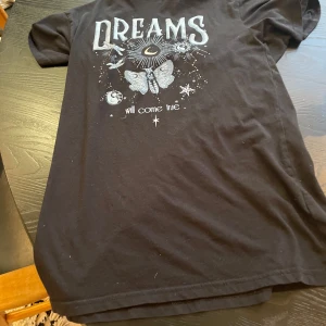 Svart t-shirt med tryck från Lindex - Svart t-shirt från Lindex med coolt vitt tryck framtill med texten 'DREAMS will come true', fjärilar, stjärnor och måne. Perfekt för dig som gillar unika prints och bekväma basplagg.