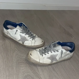 Vita sneakers från Golden Goose med grå stjärna - Säljer ett par vita sneakers från Golden Goose med grå stjärna på sidan, grå snörning och blå detalj vid hälen. Skorna har rund tå och platt sula, samt inslag av mocka på baksidan. Perfekta för en avslappnad stil.