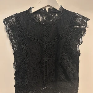 Svart spetsblus från ONLY - Säljer en svart spetsblus från ONLY med vackra detaljer och rund halsringning. Blusen är ärmlös med dekorativa volanger längs axlarna och har dragkedja i ryggen. Perfekt för dig som gillar romantisk stil.