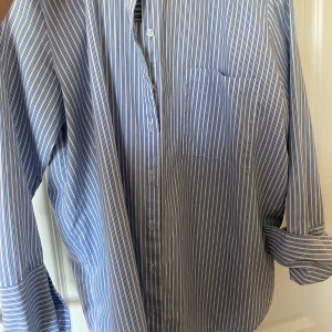 Blå och vit randig skjorta - Klassisk blå och vit randig skjorta med lång ärm och bröstficka. Skjortan har knappar framtill och en avslappnad passform, perfekt till jeans eller kostymbyxor. Jätteskönt att ha på sommaren och är i storlek M men passar som en oversized S vilket är snyggt!