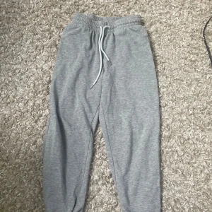 Grå mjukisbyxor med dragsko från H&M - Grå mjukisbyxor från H&M med elastisk midja och vit dragsko. Byxorna har en avslappnad passform och är perfekta för en chill vardagslook. Tillverkade i mjukt bomullsmaterial för extra komfort.