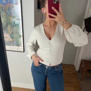 Vit blus med knappar från Gina Tricot - Säljer en stilren vit blus från Gina Tricot med långa puffärmar och knappar framtill. Blusen har en djup v-ringning och är tillverkad i ett lätt, mjukt material. Perfekt för dig som vill ha en enkel men snygg topp till garderoben.