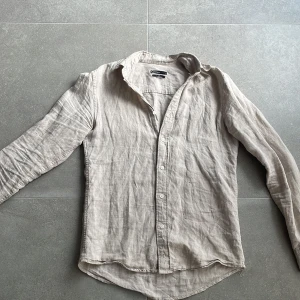Beige 100% linneskjorta  - Säljer en stilren beige skjorta i 100% linne. Skjortan har lång ärm, klassisk krage och knäppning framtill. Perfekt för en avslappnad men ändå snygg look. Regular fit-modell. Skjortan ser väldigt skrynklig ut på bilderna men eftersom det är 100% linne så blir det ofta så, inget som syns med skjortan på!