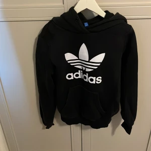 Svart hoodie från Adidas med vit logga - Svart hoodie från Adidas med stor vit logga och klassiska treklövertrycket på bröstet. Tröjan har huva och en stor magficka framtill. Perfekt för en sportig och avslappnad stil.