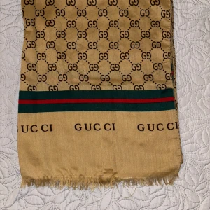 Beige halsduk från GG - Snygg beige halsduk från GG med klassiskt GG mönster och gröna samt röda ränder. Tydlig logga längs ena kanten och fransad 