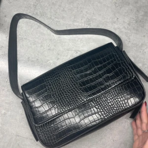 Svart axelväska i krokopräglad syntet från Bik Bok - Säljer en svart axelväska från Bik Bok i krokopräglad syntet. Väskan har magnetlås, innerfack med dragkedja och justerbar axelrem. Perfekt för att bära det viktigaste med stil.