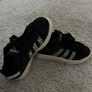 Adidas Campus sneakers med leopardmönster - Säljer ett par svarta Adidas Campus sneakers med beiga och svarta leopardmönstrade ränder på sidorna. Skorna har breda svarta snören och vit sula. Storlek 36 2/3💕använda under hösten endast så forfarande fint skick! Pris kan diskuteras!🩷
