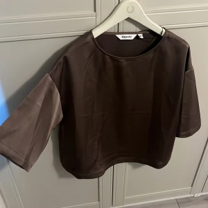 Brun croppad topp från Kappahl - Säljer en brun croppad topp från Kappahl med rund halsringning och trekvartsärmar. Toppen har en slät satinliknande yta och en enkel, stilren design som passar perfekt till många olika outfits.