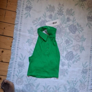 Grön ribbad polotopp från NA-KD - Säljer en grön ribbad polotopp från NA-KD med krage och knappar framtill. Toppen är ärmlös och har en tight passform, perfekt för att styla med jeans eller kjol.