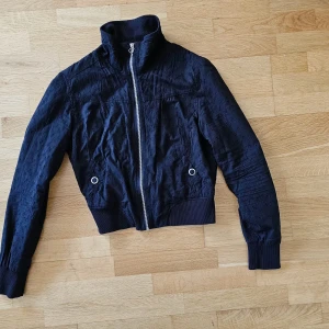 Svart jacka från Fishbone  - Svart jacka från Fishbone. Jackan har en dragkedja framtill och resår i midjan och ärmsluten. Detaljer med små hål- blommor över hela jackan. Inga skavanker! Det står stl XL på etiketten men jackan är smalare,  så passar till S och M  Jackan är gjord av bomull,  fodrad i polyester.   Längden 55 cm Bredd 52 cm mellan armhålorna  Midja 36,5 lite stretchig så går till 40 cm