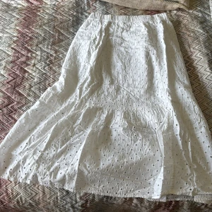 Vit broderad långkjol med volang - Säljer en vit långkjol med broderade detaljer och volang nertill. Kjolen har resår i midjan och är tillverkad i ett lätt bomullstyg med hålbroderi. Perfekt för varma dagar och ger en romantisk känsla till outfiten.