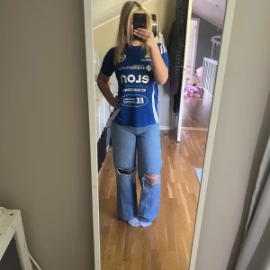 Blåa jeans med hål på knäna! - Säljer ett par blå jeans som är baggy och hål på knäna. Klassisk femficksmodell med midrise midja och knappgylf. Perfekta för en avslappnad och trendig look.