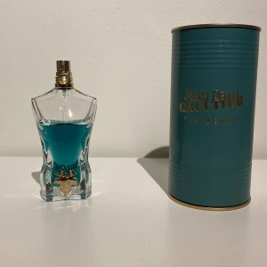 Jean Paul Gaultier Le Beau Eau de Toilette - Parfymflaska formad som en manlig torso med blått innehåll och gulddetaljer. Kommer med originalförpackning i turkos metall med guldfärgad text. En ikonisk och stilren doft för herr.  Cirka 60 ml kvar. Byten kan diskuteras.