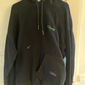 Svart hoodie från Marcelo Burlon County of Milan - Svart hoodie med broderade detaljer i grönt, rött och lila från Marcelo Burlon County of Milan. Tröjan har huva med dragsko och en stor magficka framtill. Perfekt för en avslappnad och trendig stil. Storlek M oversized men väldigt snygg på 