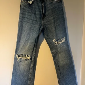 Blå slitna jeans från Zara - Säljer ett par blå jeans från Zara med raka ben och slitna detaljer på båda knäna. Jeansen har normal passform med en hög midja. Storlek 38, lite mindre i storleken. 