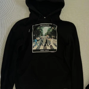 Svart hoodie med The Beatles Abbey Road-tryck - Svart hoodie med tryck av The Beatles Abbey Road-albumomslag på bröstet. Tröjan har huva med dragsko och en klassisk känguruficka framtill. 